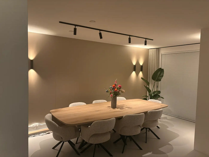 Zwarte railspots op een rail boven een eikenhouten eettafel, met zwarte wandlampen die de wand in de avond verlichten
